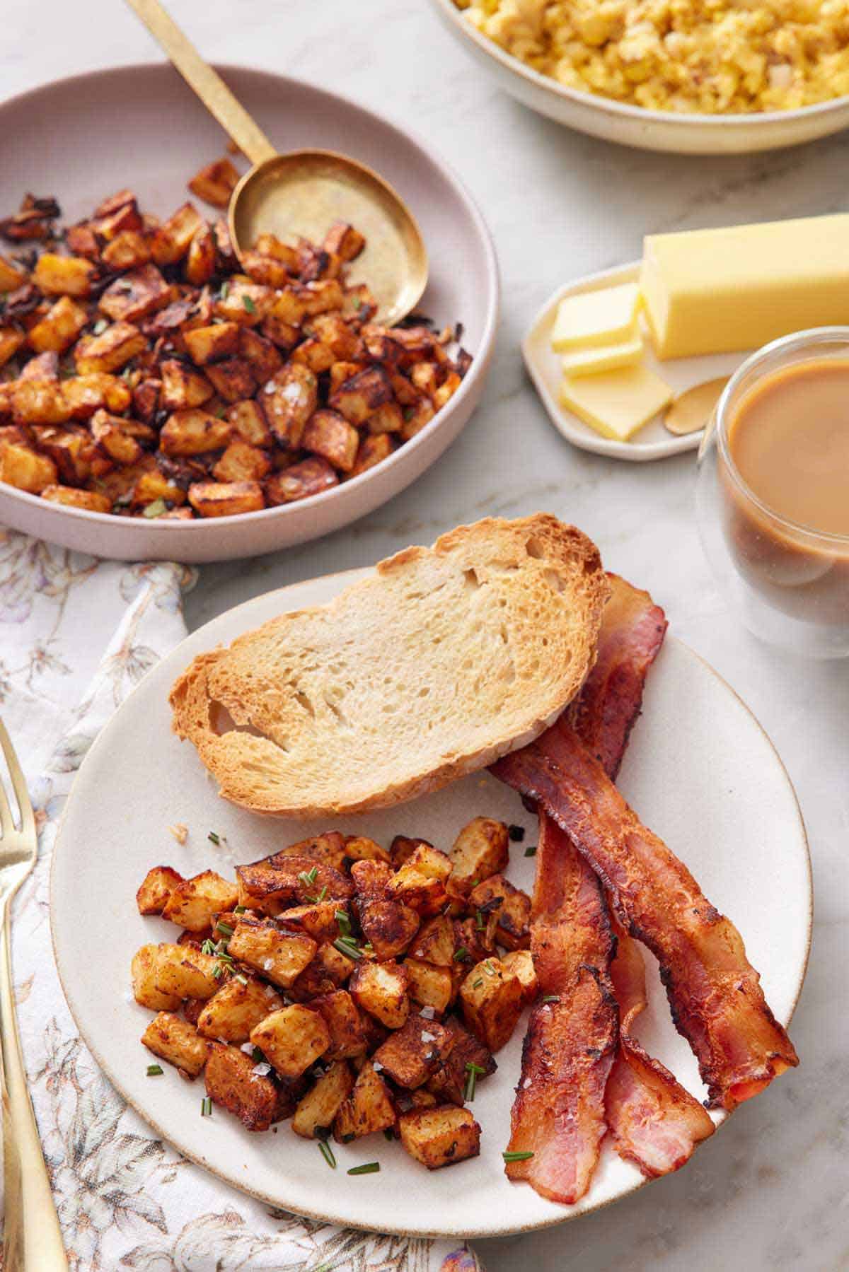 A plate with breakfast potatoes, bacon strips, and toast. A plate with more breakfast potatoes, a cup of coffee, and butter in the background.