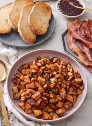 A plate with breakfast potatoes topped with chopped rosemary. A plate of toast and bacon in the back.