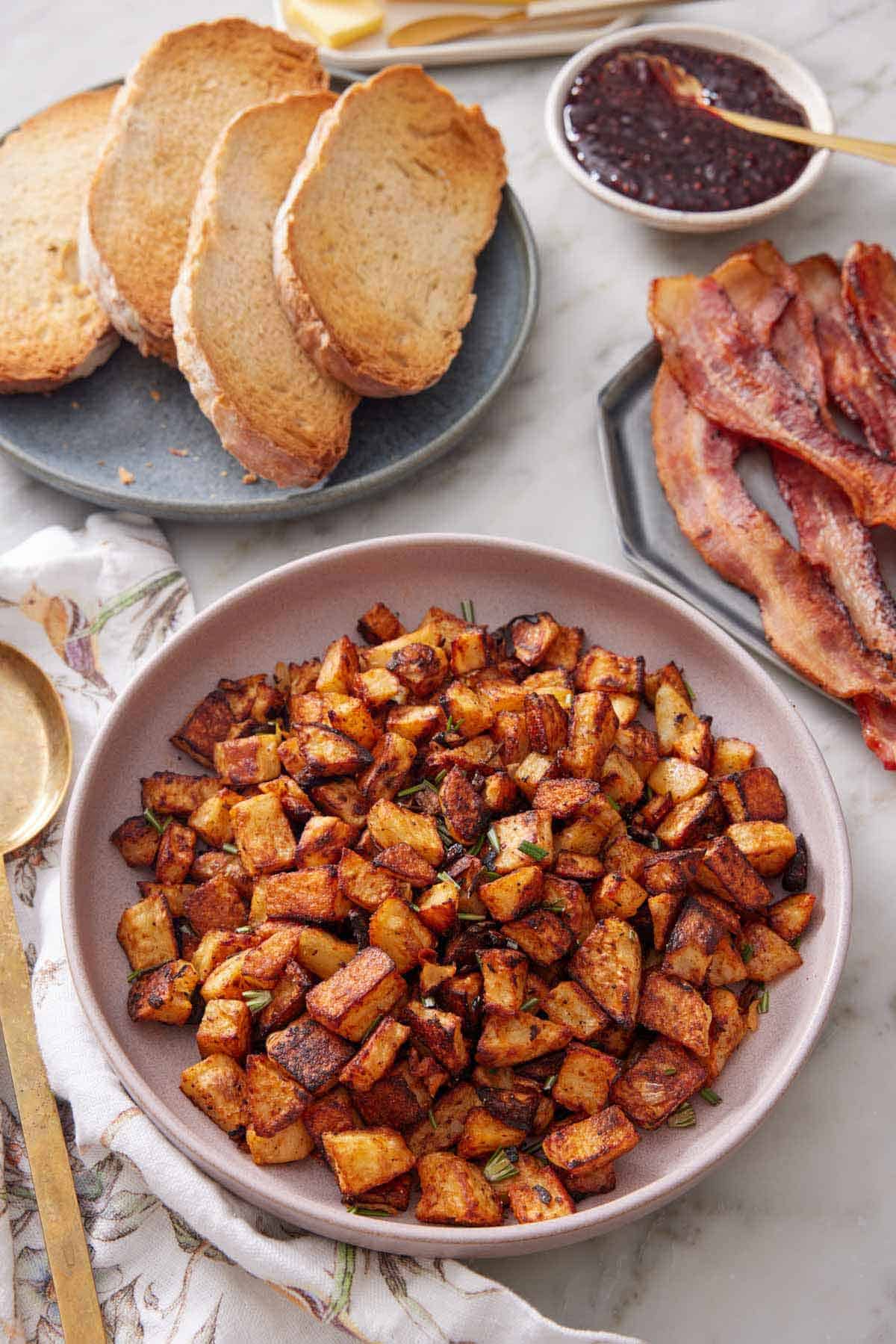 A plate with breakfast potatoes topped with chopped rosemary. A plate of toast and bacon in the back.