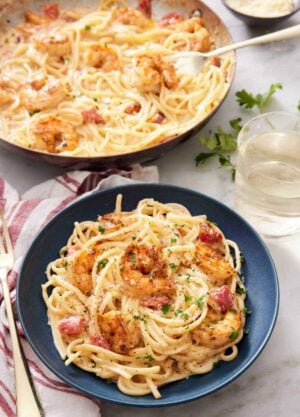 A plate of cajun shrimp pasta with chopped parsley garnish with a skillet with more pasta in the background. A glass of win on the side.