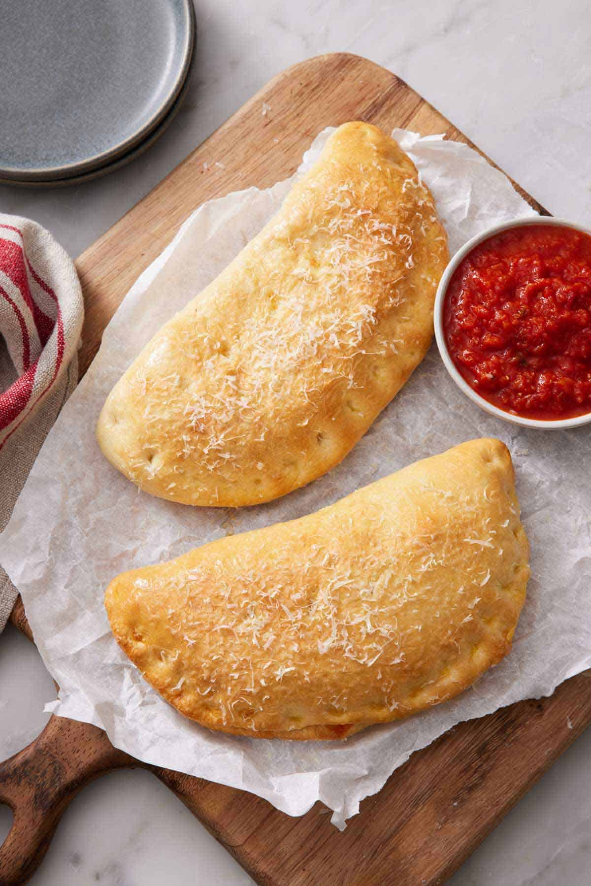 Overhead view of two calzones on a sheet of parchment over a wooden serving board with a bowl of pizza sauce.