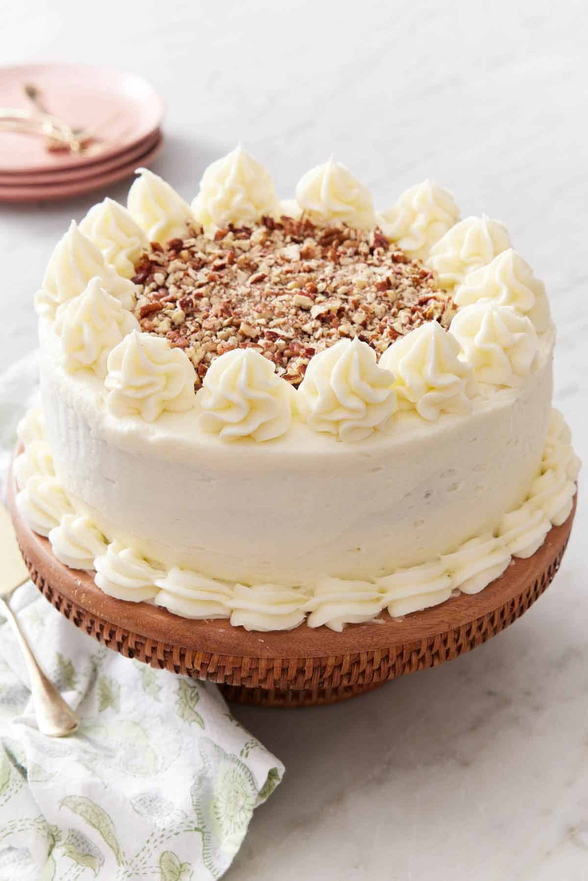 A carrot cake coated in frosting and topped with chopped pecans on a cake stand.