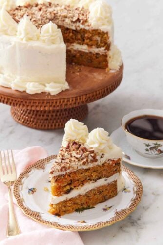 A slice of carrot cake with the rest of the cake on a cake stand in the back along with a cup of coffee.