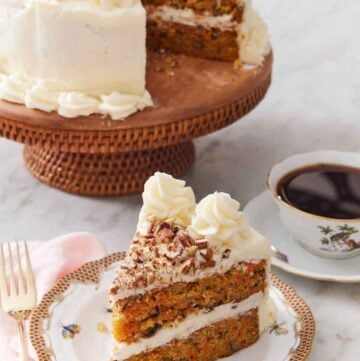 A slice of carrot cake with the rest of the cake on a cake stand in the back along with a cup of coffee.