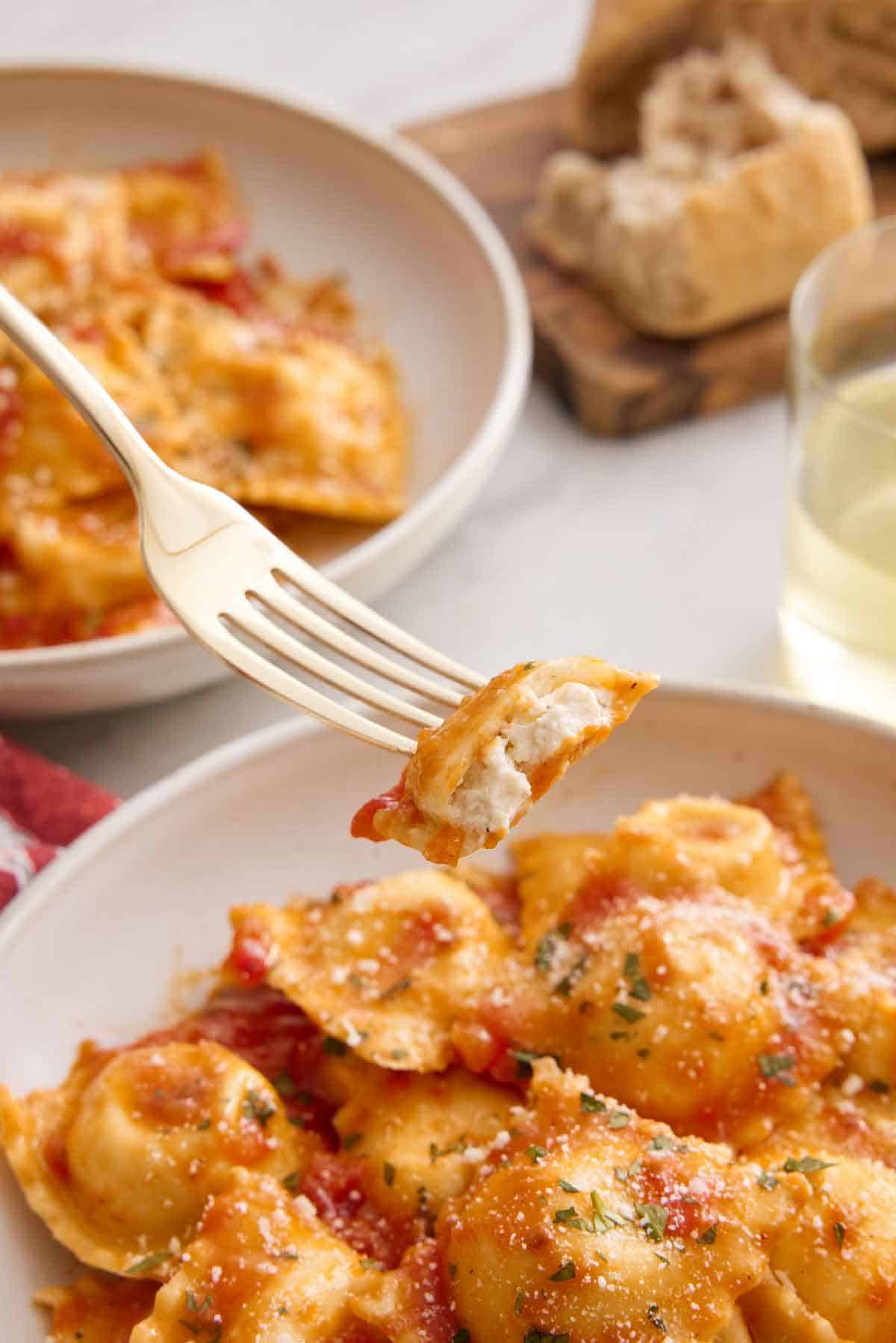 A fork lifting up a bite of cheese ravioli tossed in tomato sauce and herb garnish. A second plate, torn bread, and wine in the background.