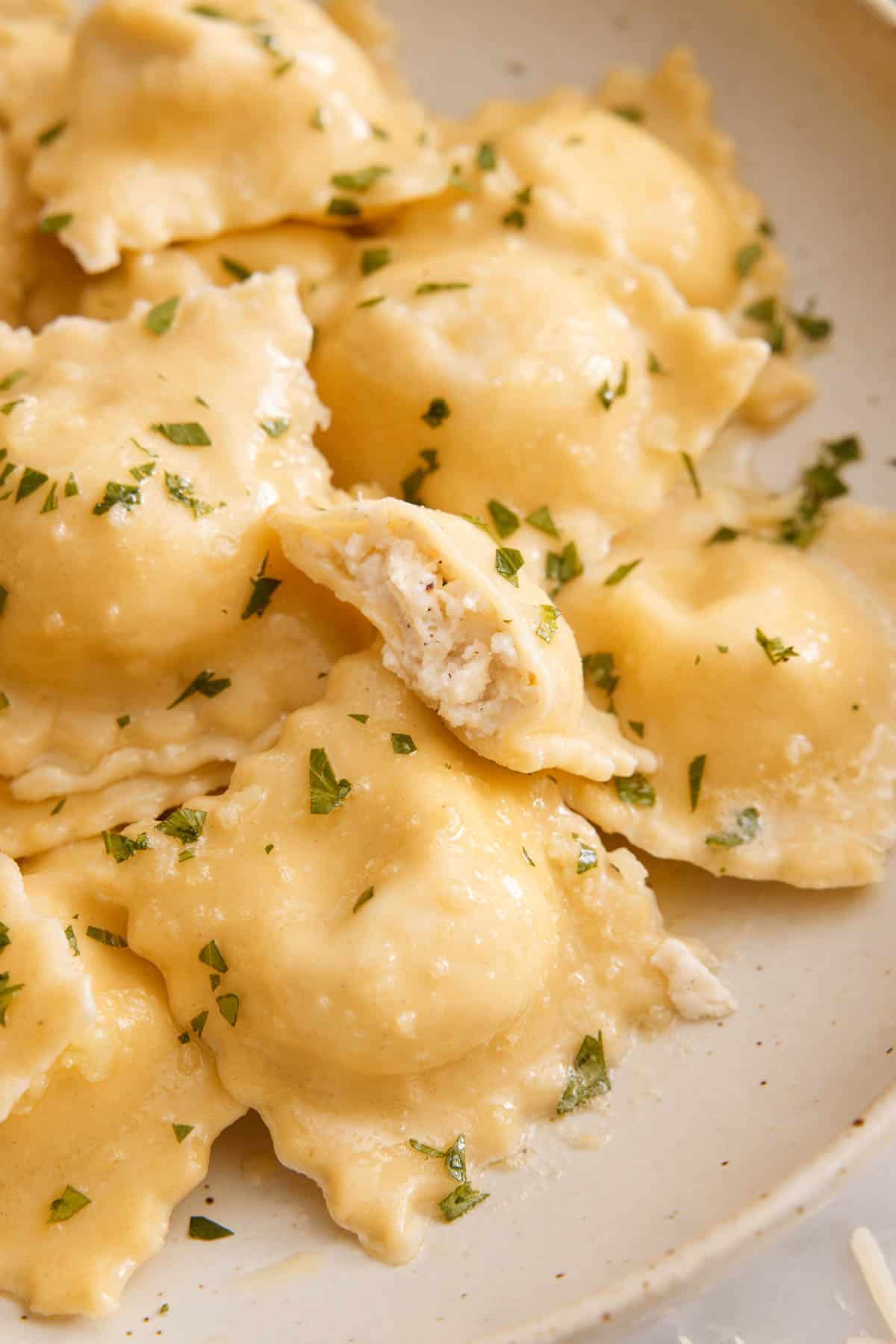 A close up view of a plate of cheese ravioli garnished with chopped parsley. One ravioli bitten in half, showing the cheese filling.