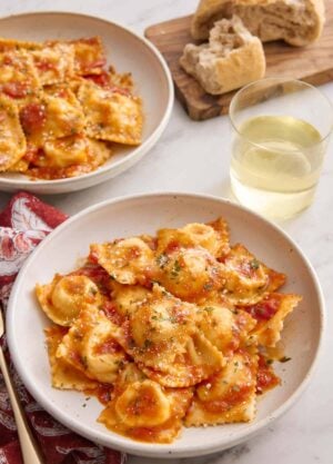 A plate of cheese ravioli tossed in tomato sauce with a glass of wine, torn bread, and another plate of ravioli in the background.