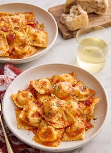 A plate of cheese ravioli tossed in tomato sauce with a glass of wine, torn bread, and another plate of ravioli in the background.