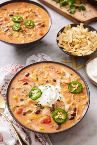 Two bowls of chicken enchilada soup topped with sliced jalapeno and one bowl with sour cream. A bowl of tortilla strips on the side.