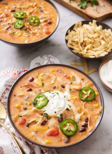 Two bowls of chicken enchilada soup topped with sliced jalapeno and one bowl with sour cream. A bowl of tortilla strips on the side.