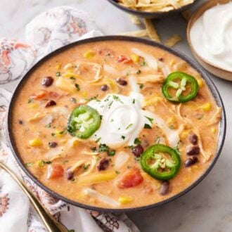 A bowl of chicken enchilada soup with soup cream and sliced jalapenos. A bowl of sour cream in the background and a spoon beside the soup.