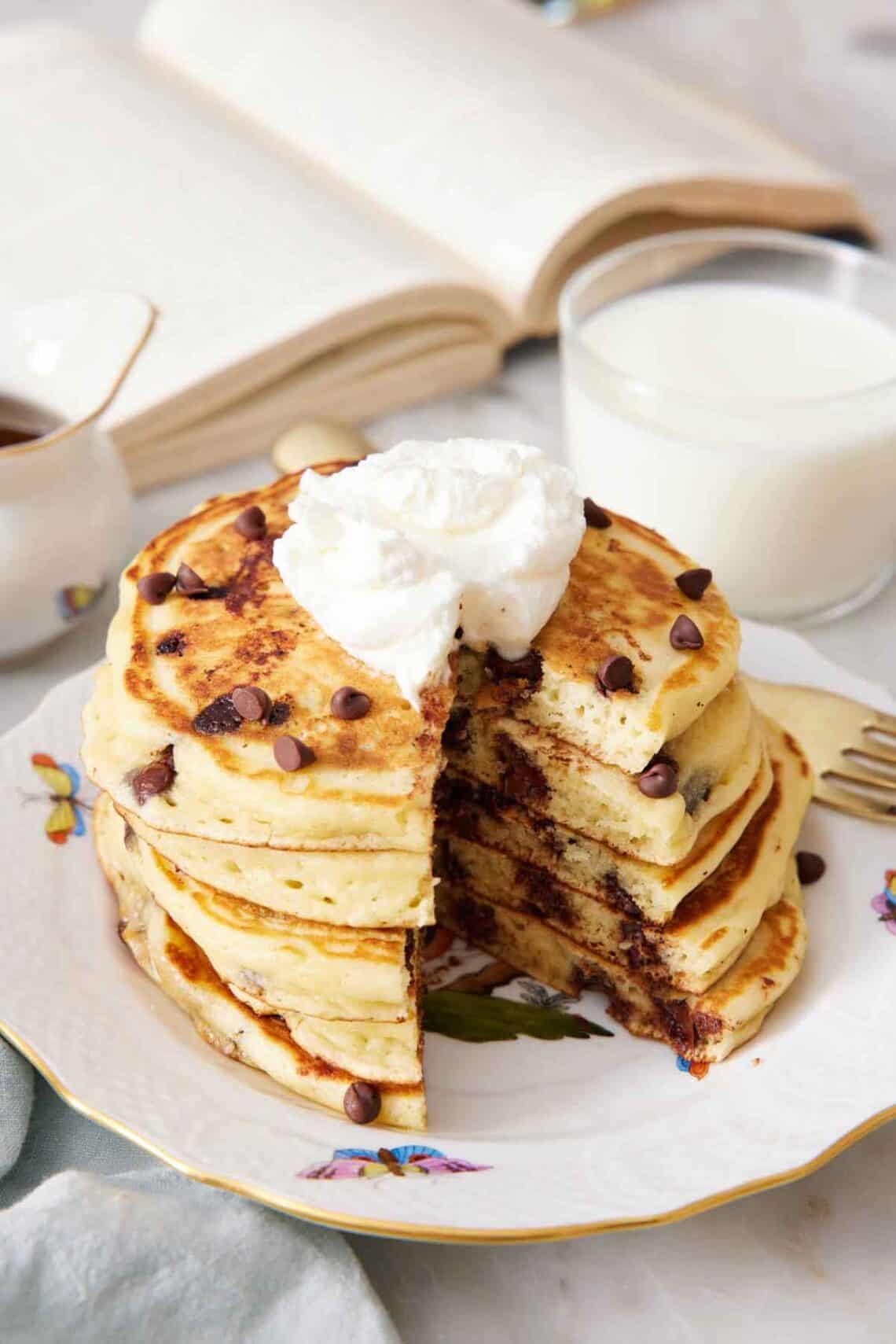 A stack of cut chocolate chip pancakes with whipped cream on top with a glass of milk and opened book in the back.