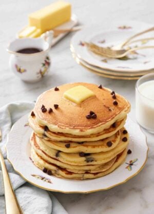 A stack of chocolate chip pancakes with a pat of butter on top with a few extra chocolate chips. A stack of plates and forks in the background along with syrup, butter, and milk.