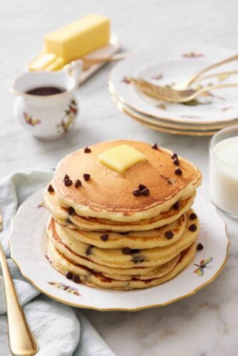 A stack of chocolate chip pancakes with a pat of butter on top with a few extra chocolate chips. A stack of plates and forks in the background along with syrup, butter, and milk.