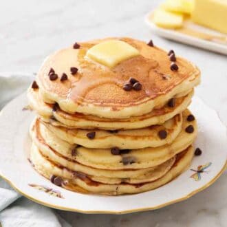 A stack of chocolate chip pancakes on a plate with extra chocolate chips, butter, and syrup on top. Butter in the background.
