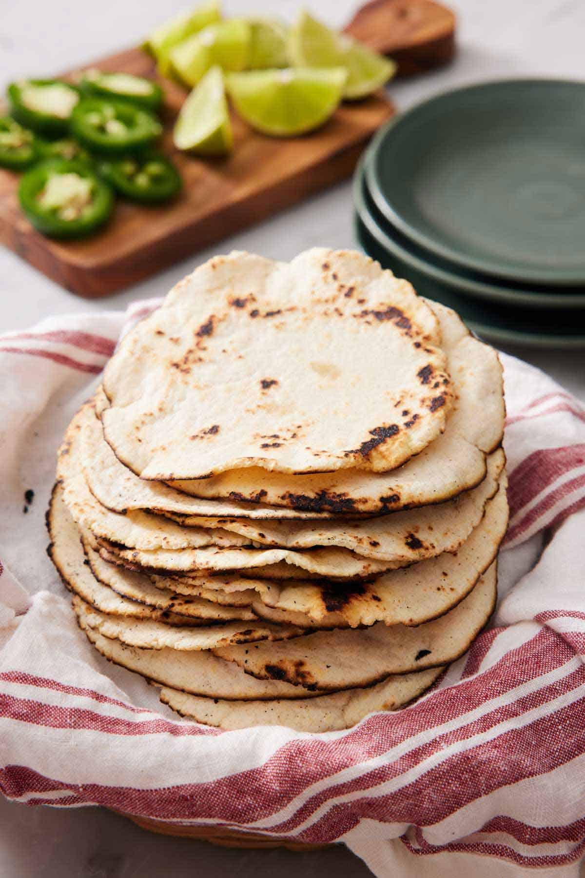 A stack of corn tortillas on a linen napkin with a stack of plate in the background along with sliced jalapenos and limes.