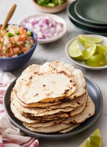A stack of corn tortillas on a plate. A plate of limes, plate of diced onions, bowl of salsa, and bowl of jalapeno in the background along with a stack of plates.