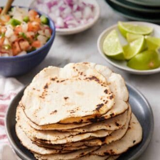 Pinterest graphic of a stack of corn tortillas on a plate. Limes, diced onions, salsa, and jalapenos in the background along with a stack of plates.