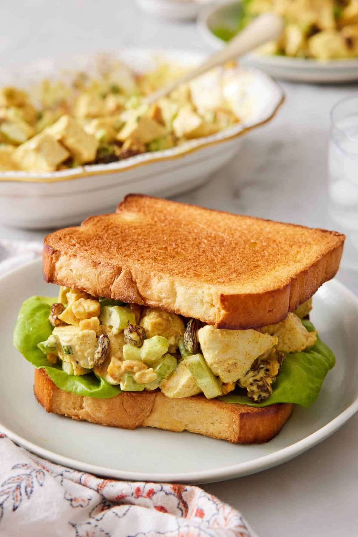 A plate with curried chicken salad on lettuce between two toasted pieces of bread. A platter of curried chicken salad in the back.