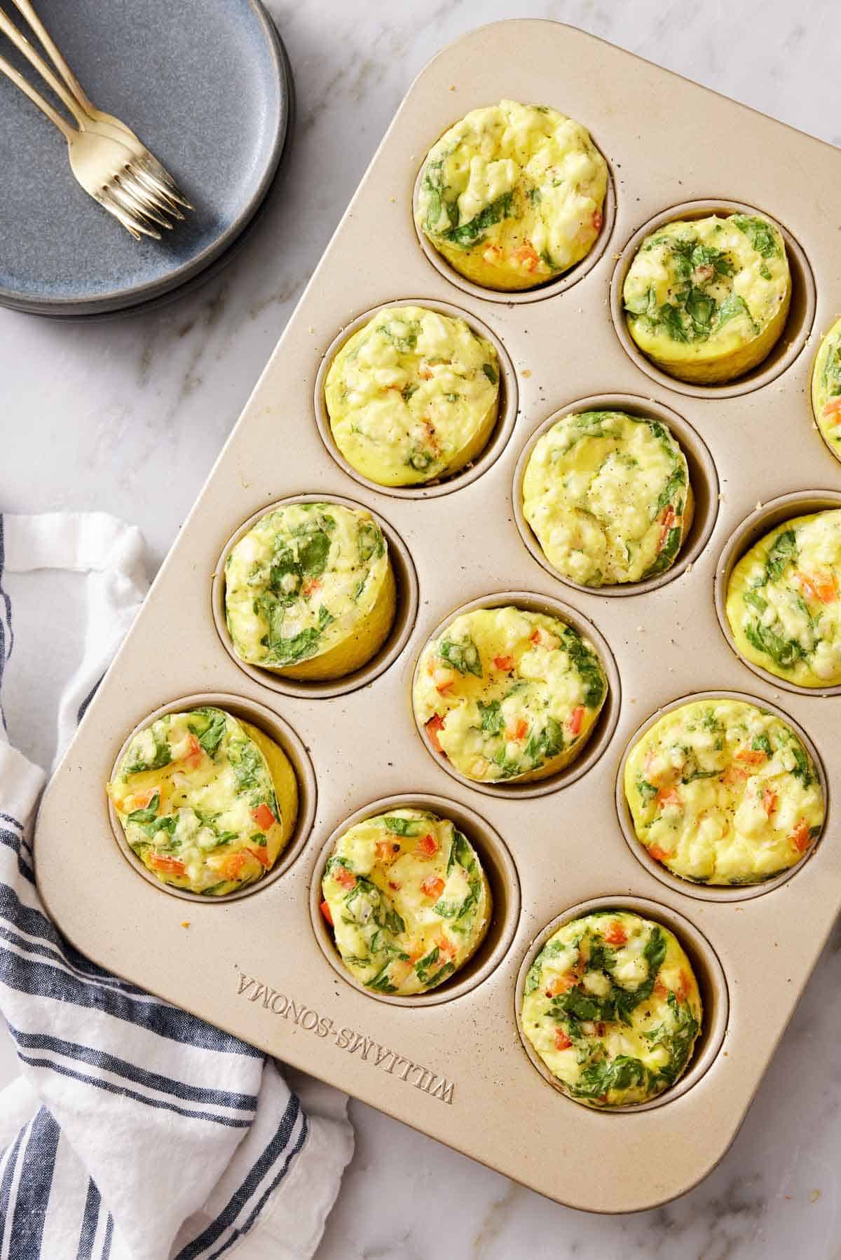 Overhead view of egg muffins in a muffin tin. A linen napkin, stack of plates, and forks off to the side.