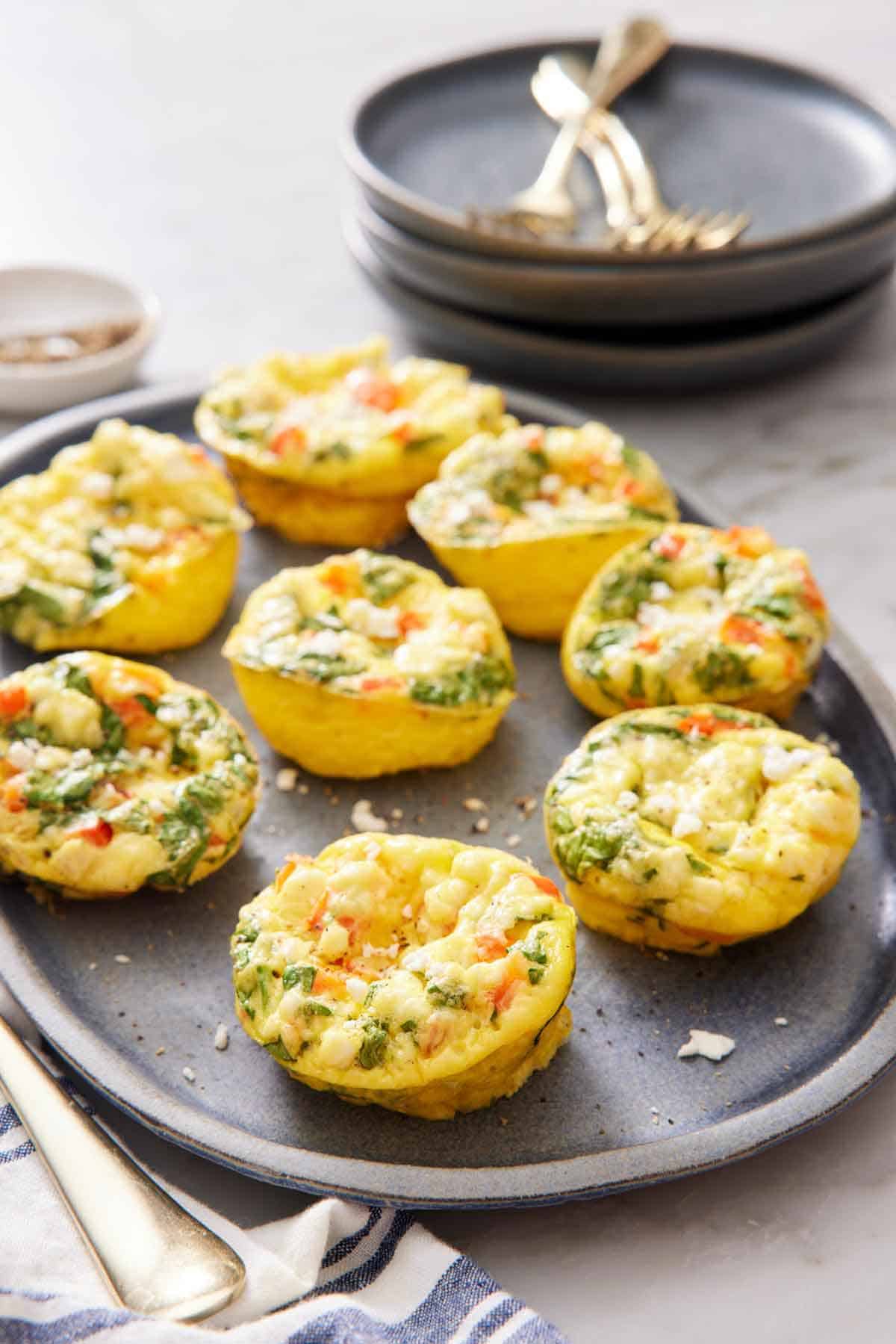 A platter of egg muffins topped with crumbled feta. Stack of plates and forks in the background.