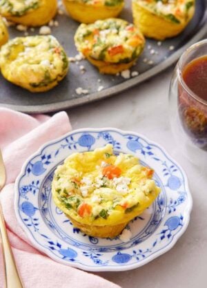 A plate with an egg muffin topped with crumbled feta cheese. A platter with more egg muffins and a coffee in the background.