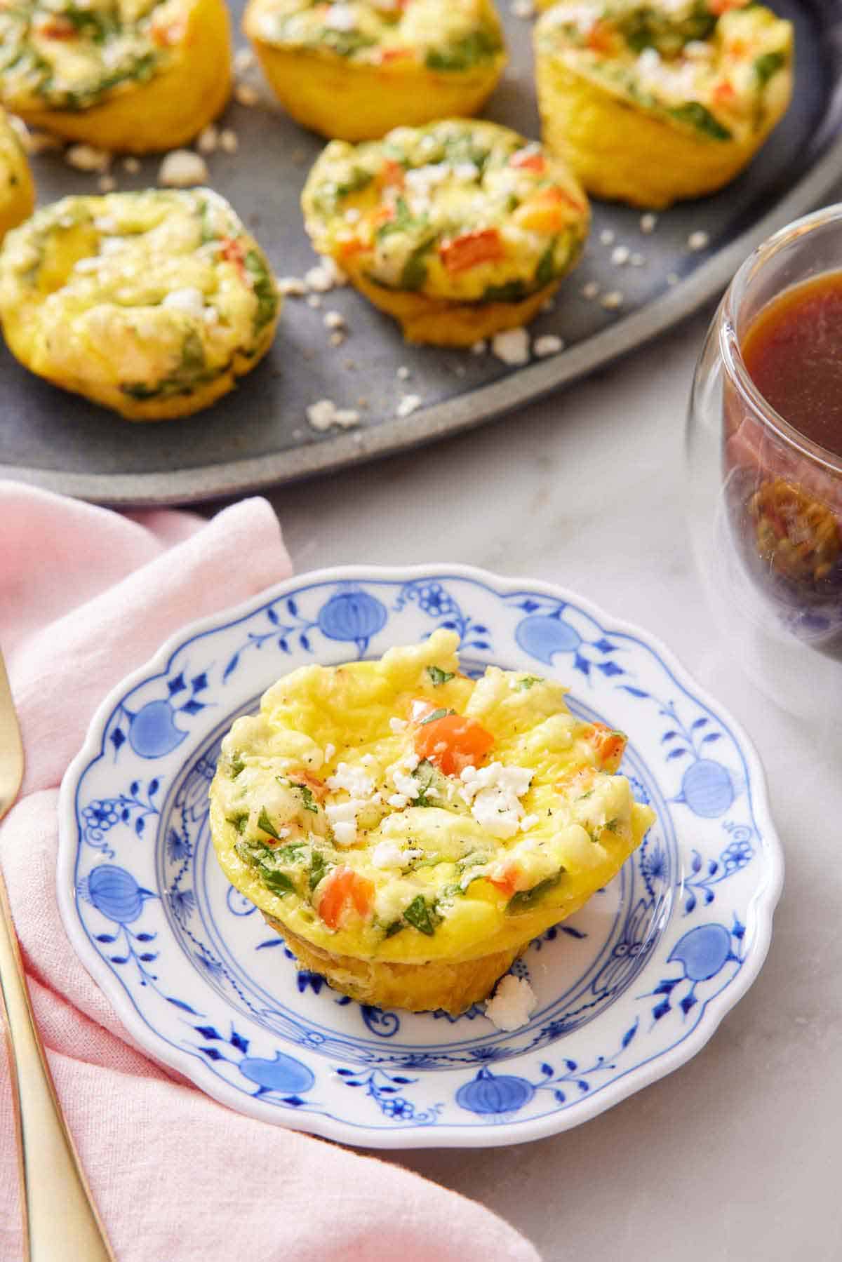 A plate with an egg muffin topped with crumbled feta cheese. A platter with more egg muffins and a coffee in the background.