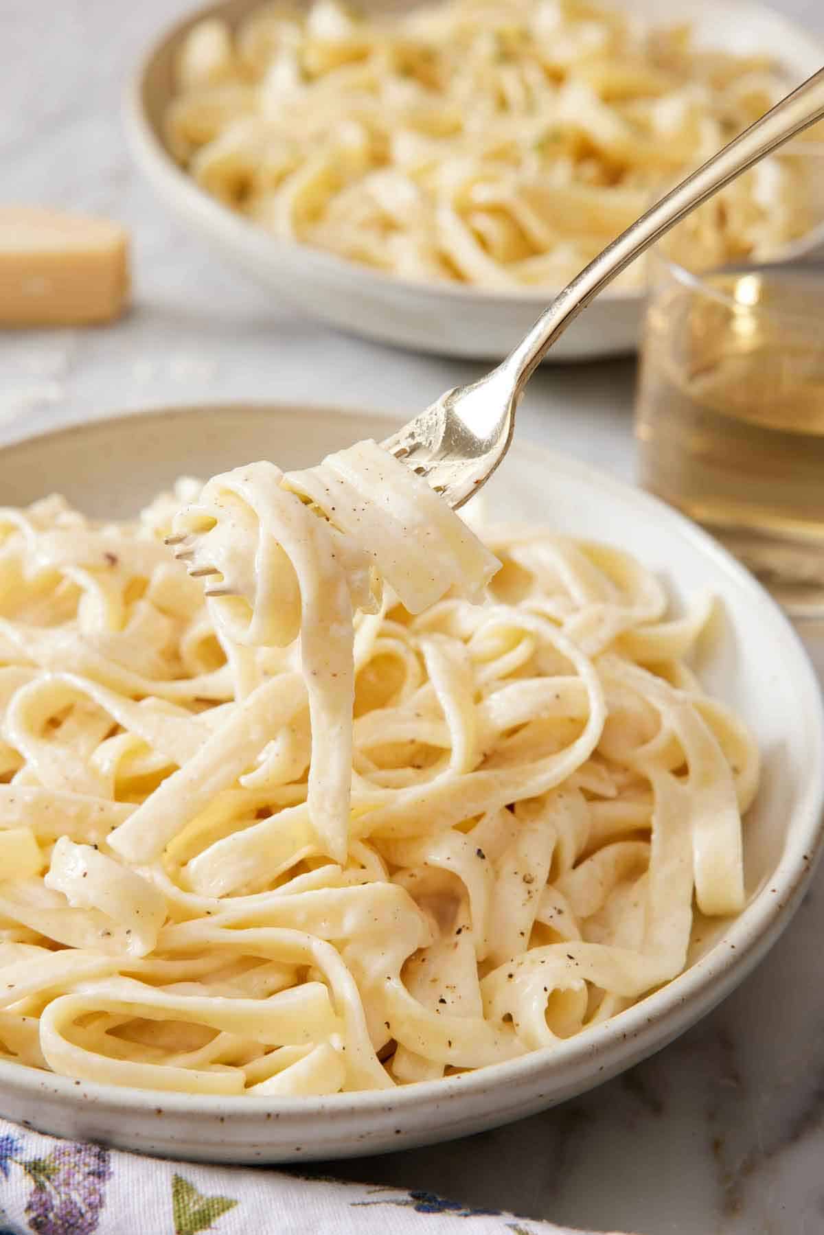 A fork lifting up a bite of fettucine alfredo from a plate. A second plate and glass of wine in the background.