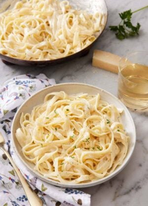 A bowl of fettucine alfredo garnished with chopped parsley with a glass of wine and a skillet with more pasta in the back.