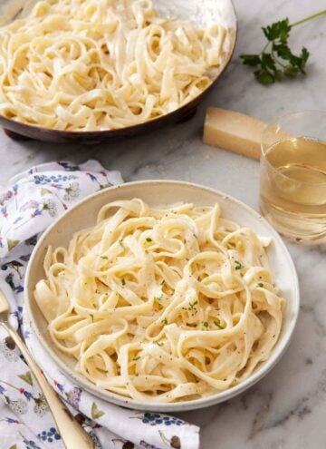 A bowl of fettucine alfredo garnished with chopped parsley with a glass of wine and a skillet with more pasta in the back.