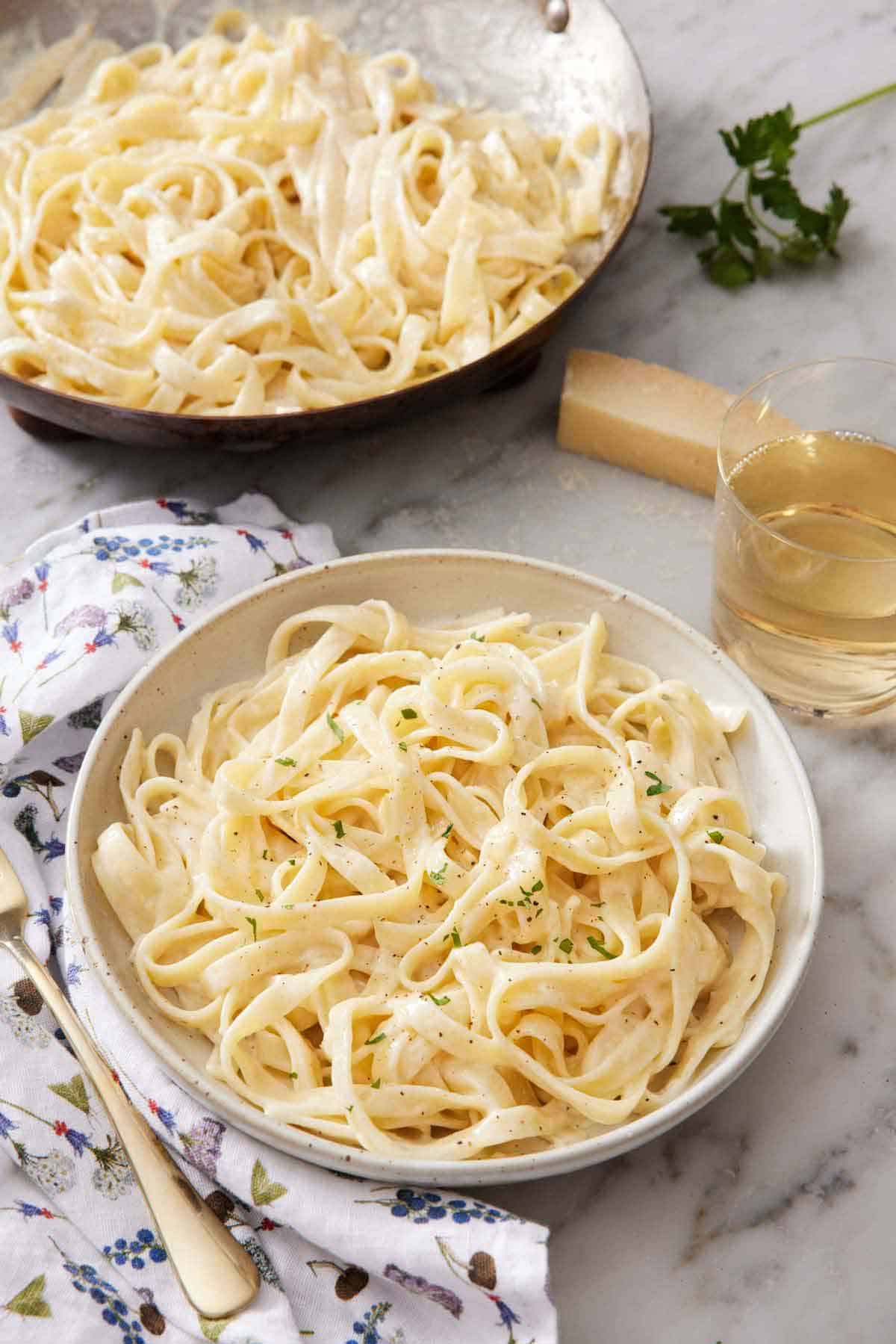A bowl of fettucine alfredo garnished with chopped parsley with a glass of wine and a skillet with more pasta in the back.