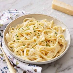 A plate of fettucine alfredo with a fork beside it on a napkin with parmesan in the background.