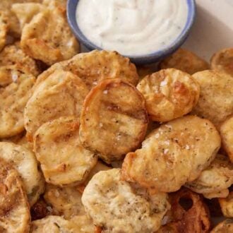 A close up of fried pickles in a platter topped with flaky salt. Bowl of sauce in the platter off to the side.