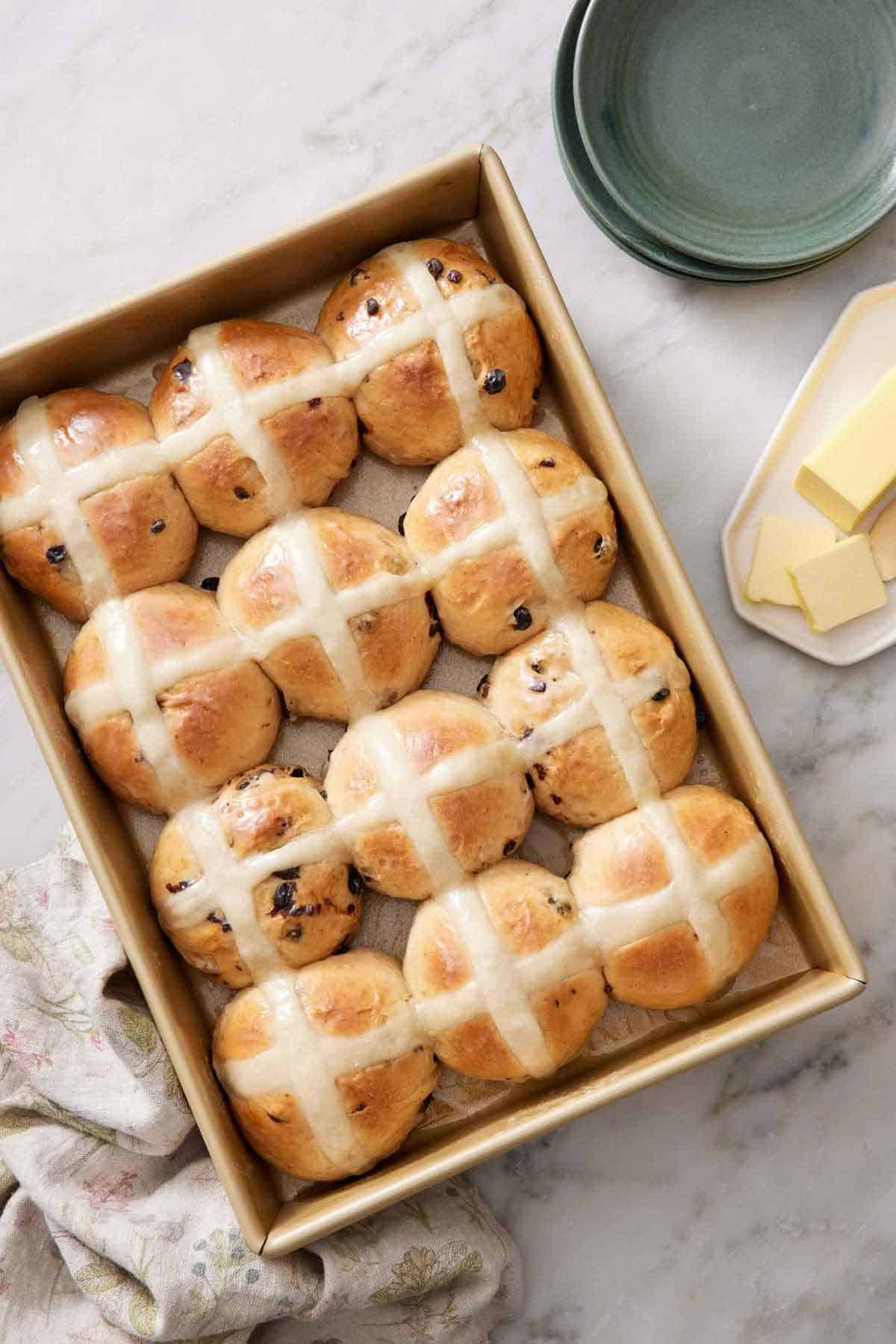 Overhead view of hot cross buns in a baking dish. Plates and butter on the side.