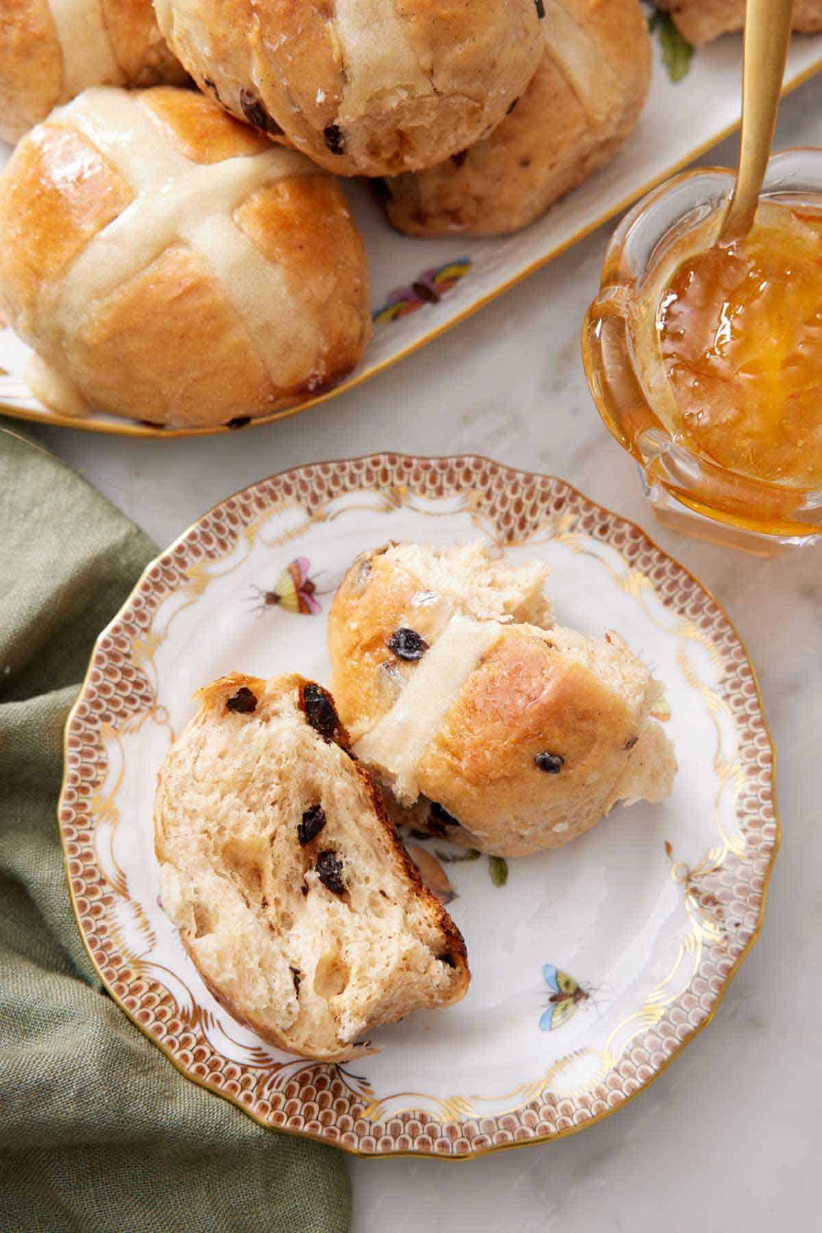 A plate with a hot cross bun torn in half. A platter of more buns and some jam off to the back.