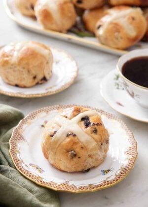 A plate with a hot cross bun with a second plate in the back along with a platter and a cup of coffee.