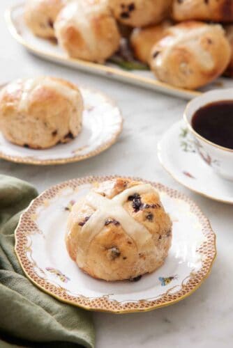 A plate with a hot cross bun with a second plate in the back along with a platter and a cup of coffee.
