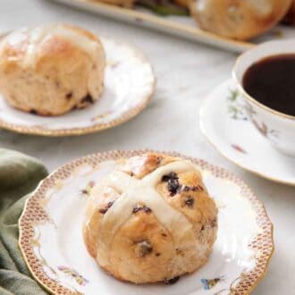Pinterest graphic of a plate with a hot cross bun with a second plate in the back along with a platter and a cup of coffee.