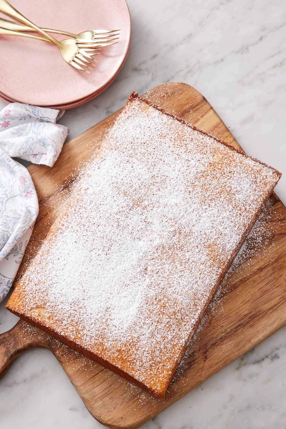 Overhead view of an uncut slab of hot milk cake dusted with powdered sugar on a wooden serving board. Plates and forks on the side.