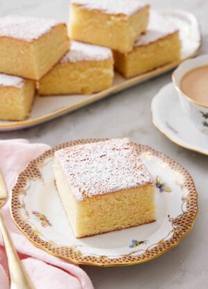 A slice of hot milk cake topped with a dusting of powdered sugar. A platter with more pieces in the back.