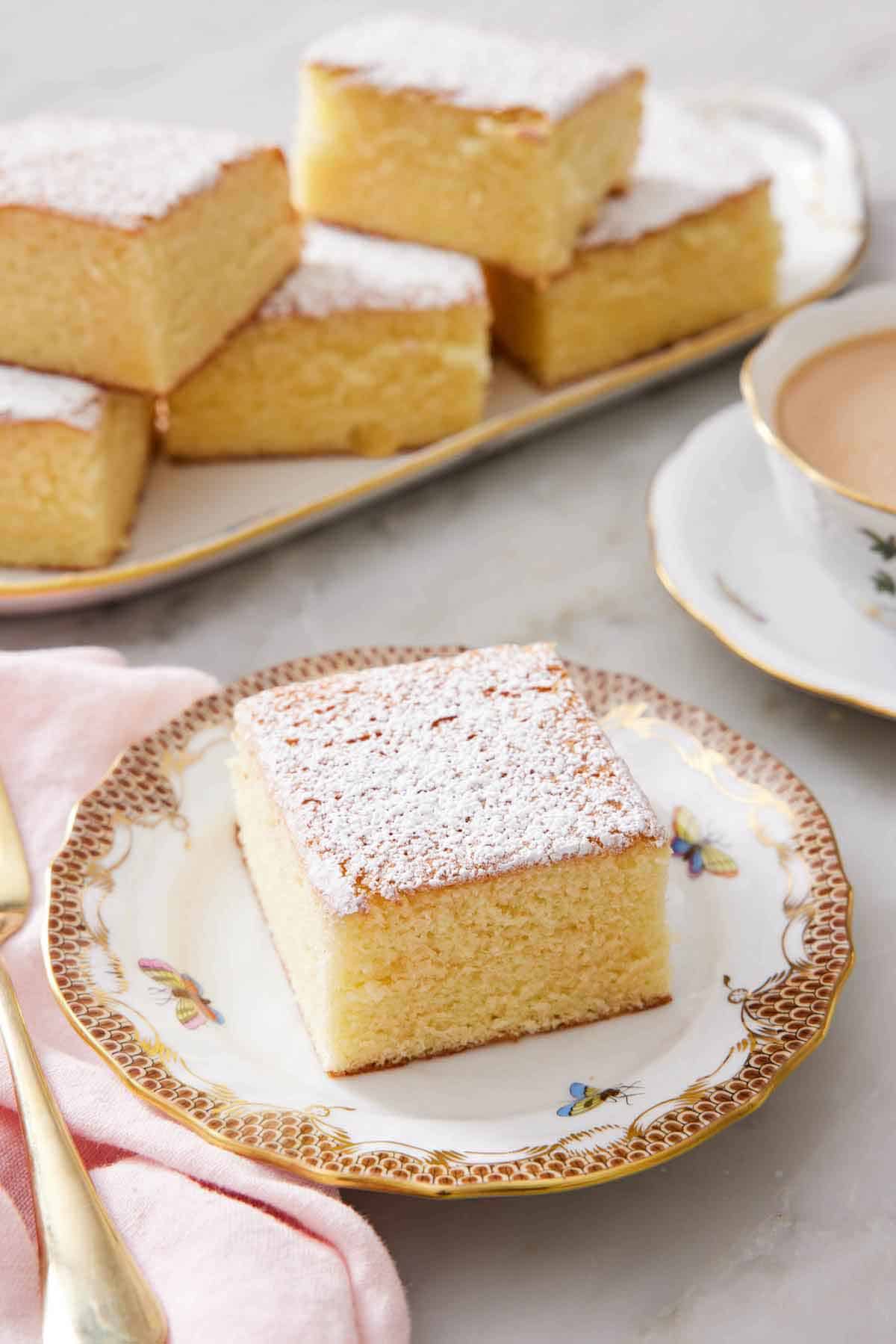 A slice of hot milk cake topped with a dusting of powdered sugar. A platter with more pieces in the back.