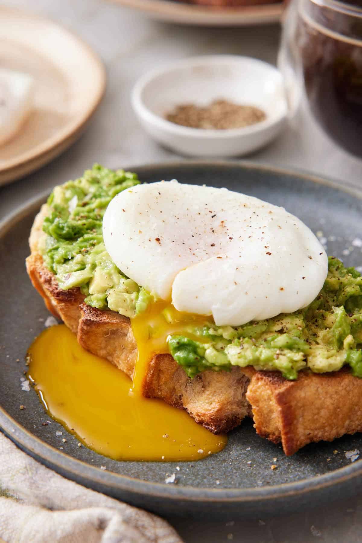 A plate with avocado toast topped with a poached egg with the yolk dripping down the bread. A glass of coffee and a small bowl of pepper in the back.