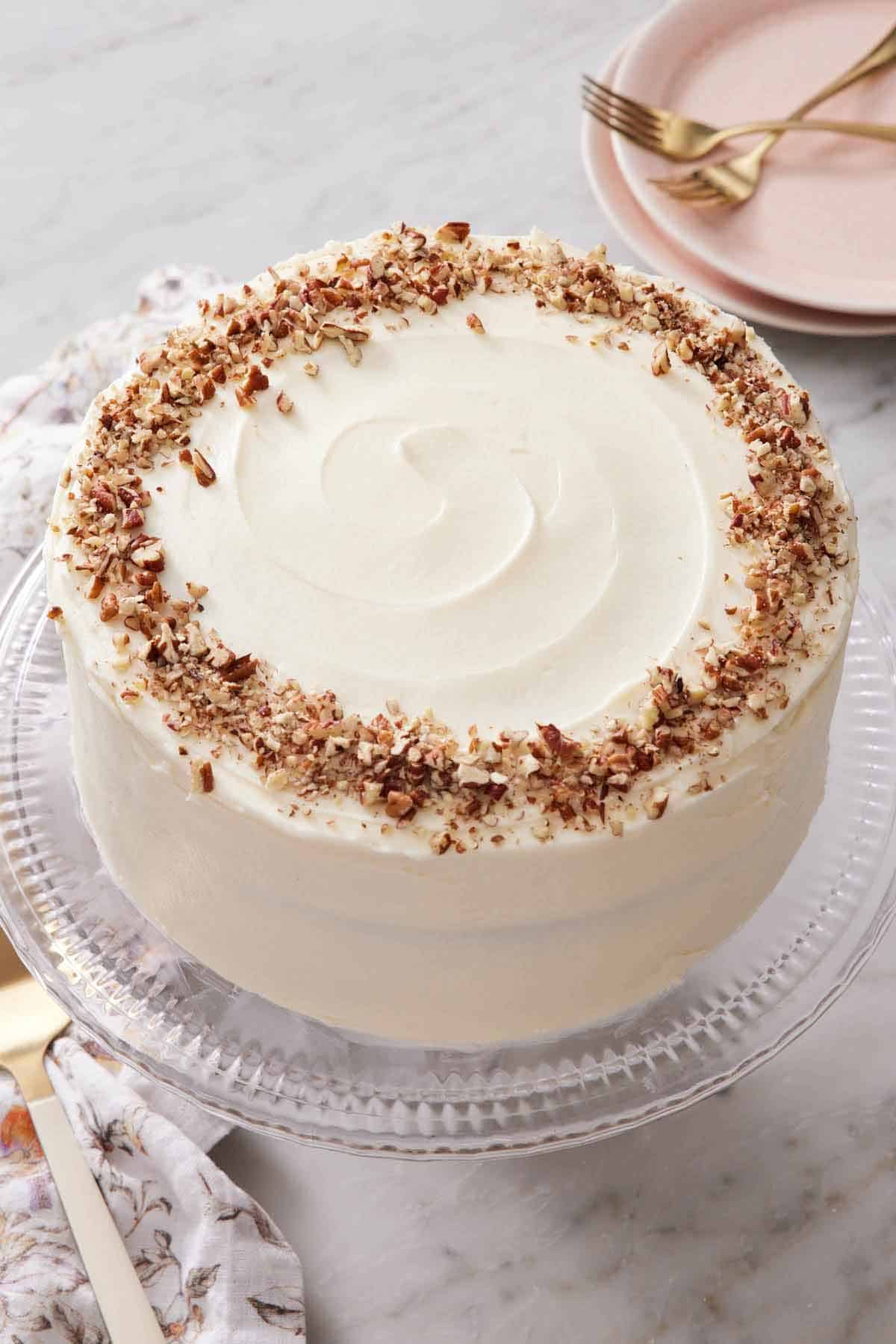 A clear cake stand with a hummingbird cake topped with chopped pecans.