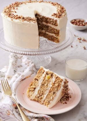 A slice of hummingbird cake on a plate with a cake stand with the rest of the cake in the background along with a glass of milk.