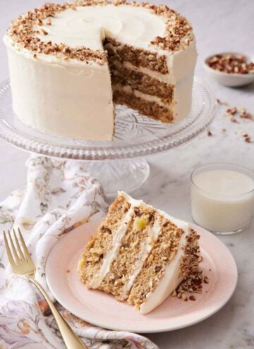A slice of hummingbird cake on a plate with a cake stand with the rest of the cake in the background along with a glass of milk.