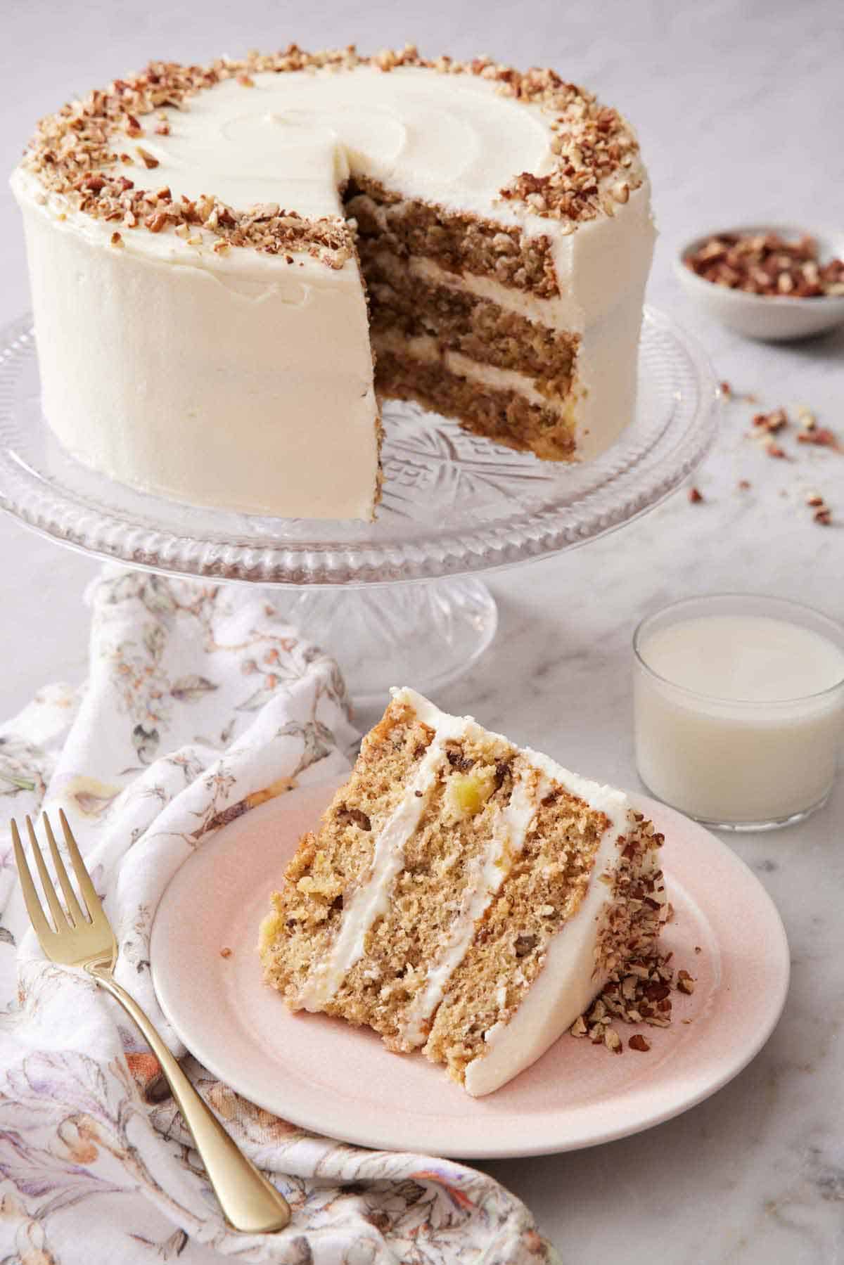 A slice of hummingbird cake on a plate with a cake stand with the rest of the cake in the background along with a glass of milk.