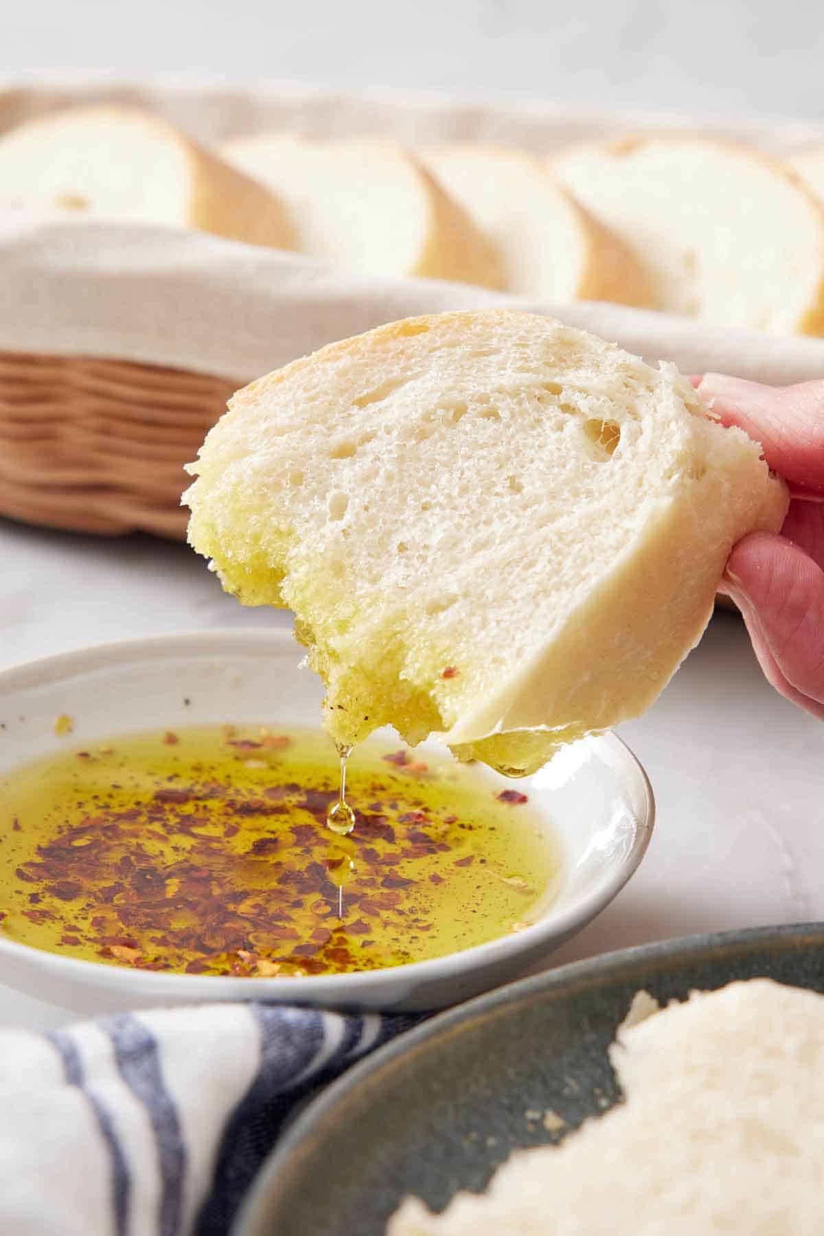 A piece of Italian bread dipped into oil with a basket with more bread in the background.