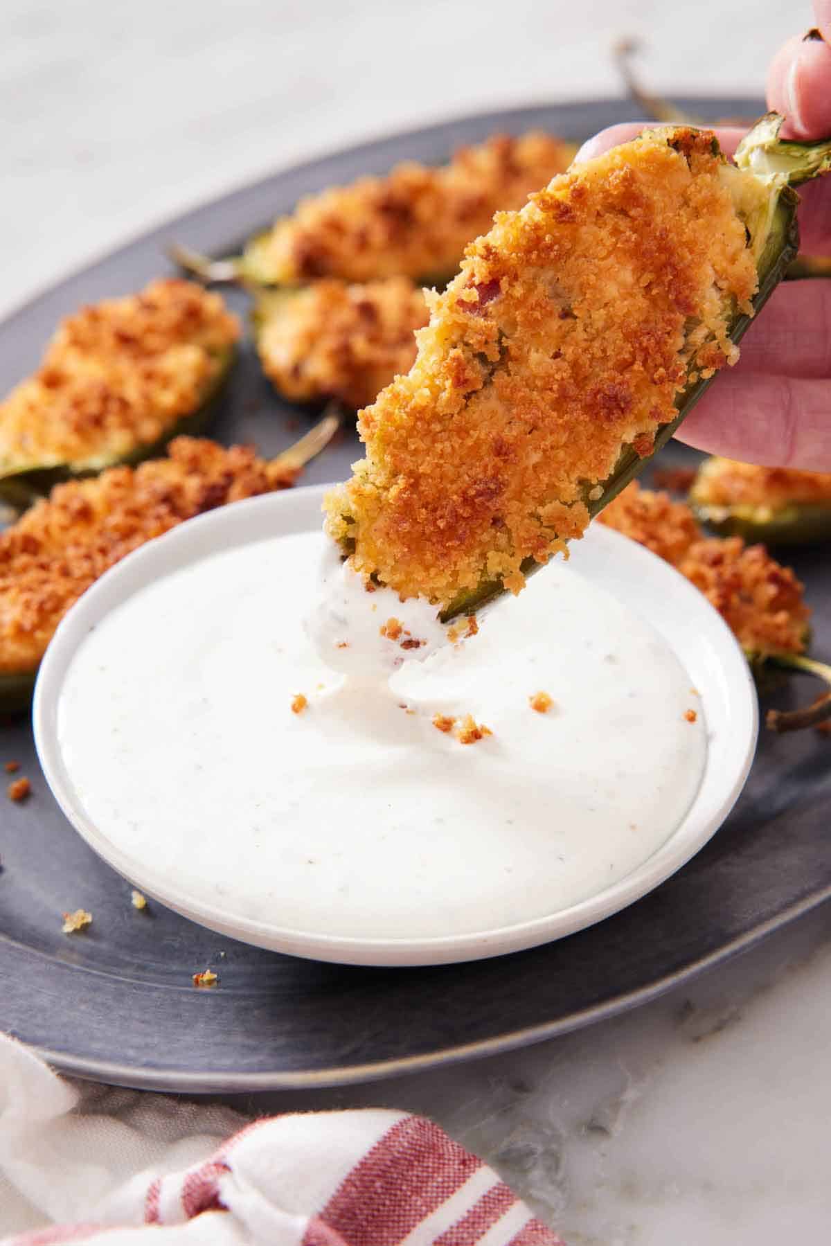 A jalapeno popper dipped into a creamy dipping sauce with more jalapeno poppers on the platter.