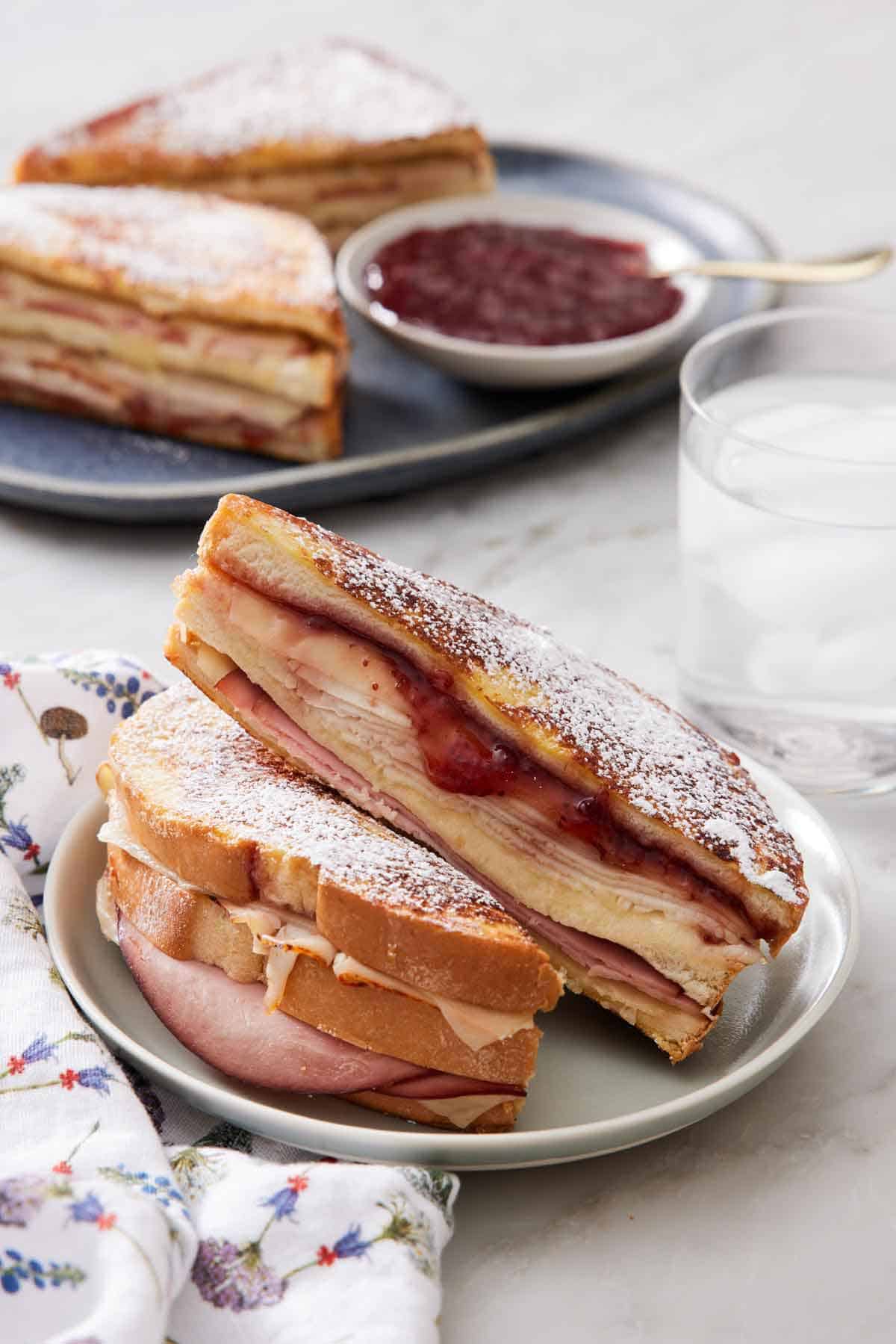 A plate with a Monte Cristo sandwich cut in half with another one in the background along with a glass of water.