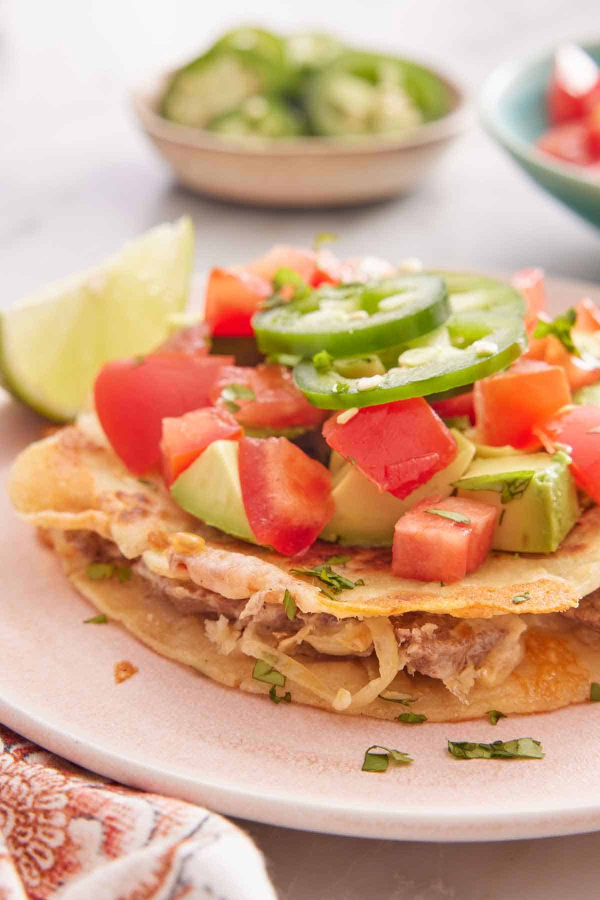 A close profile view of a plate with mulitas topped with diced tomatoes, avocados, and jalapenos with a lime beside it.
